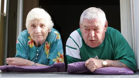 Ein älteres Ehepaar schaut, auf Kissen gelehnt,  aus dem Fenster