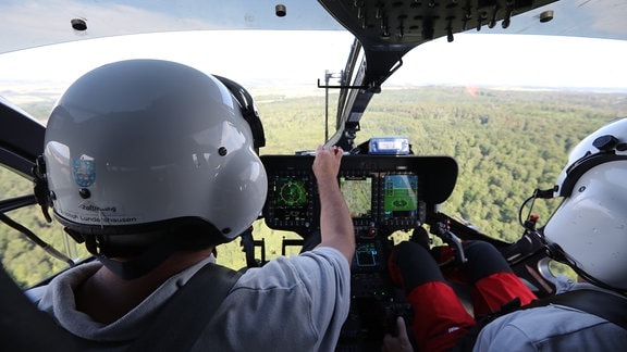 Blick aus der Kabine eines Hubschraubers, im Vordergrund die Helme zweier Piloten.