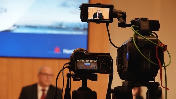 Blick auf eine Fernsehkamera, die auf Ties Rabe ausgerichtet ist, der an einem Tisch im Raum der Landespressekonferenz (LPK) sitzt.