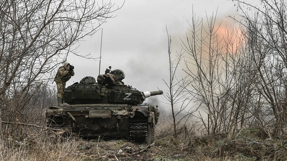 Ein Panzer schießt während sich ein darauf stehender Soldat die Ohren zu hält.