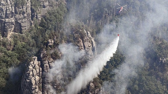 Ein Lastenhubschrauber aus Österreich fliegt mit einem Löschwasser-Außenlastbehälter um einen Waldbrand im Nationalpark Sächsische Schweiz zu löschen.