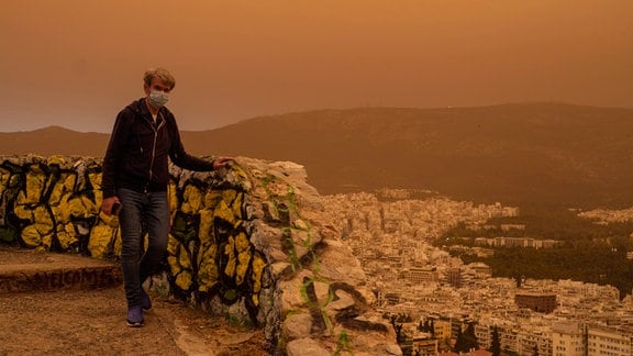Ein Mann mit einer Maske geht auf dem Lycabettus-Hügel spazieren, während im Hintergrund die Stadt Athen zu sehen ist. 