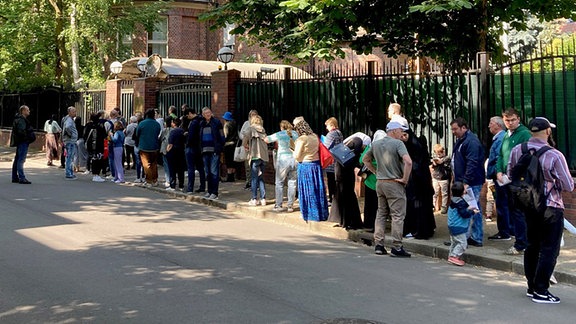 Menschen stehen vor dem Eingang einer backsteinfarbenen Villa an. 