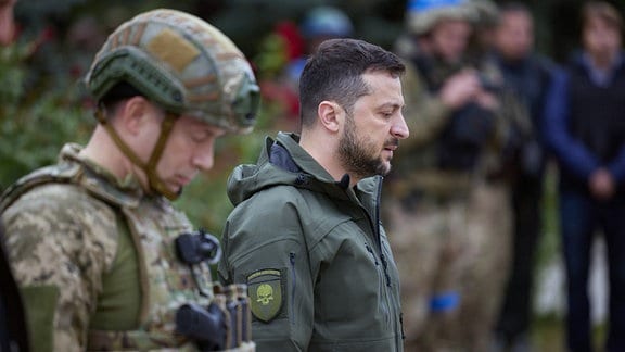 Wolodymyr Selenskyj steht neben einen Soldaten, beide mit gesenkten Köpfen. Auch bei diesem Besuch in Isjum am 14. September 2022 trägt er ein Militärabzeichen mit einem Totenkopfabzeichen. 
