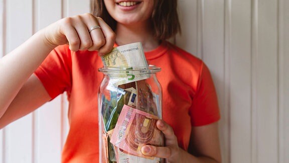 Mädchen steckt einen Fünf-Euro-Schein in ein Einweckglas mit Geldscheinen.