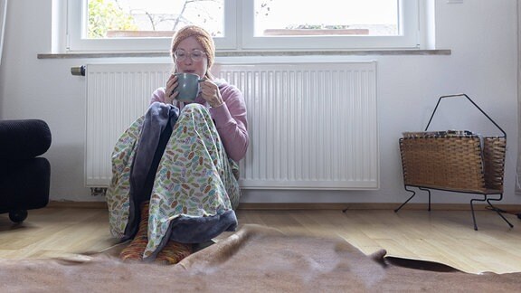 Frau sitzt, in eine Decke gewickelt, vor einem Heizkörper und bläst in eine Tasse.