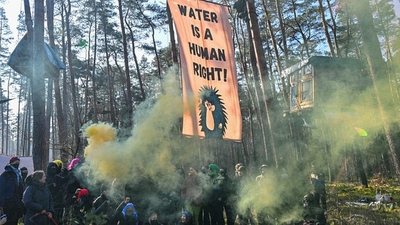 Aktivisten der Initiative "Tesla stoppen" stehen mit Rauchfackeln vor einem Transparent mit der Aufschrift "Water is a human right!" in einem Kiefernwald nahe dem Werk der Tesla-Gigafactory Berlin-Brandenburg.