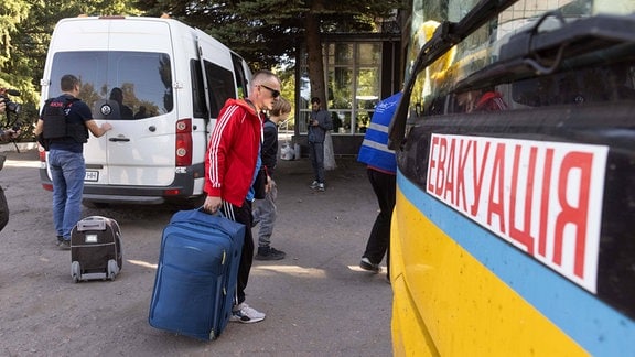 Menschen mit Koffern laufen an einem Transporter mit der Aufschrift "Evakuierung" vorbei.