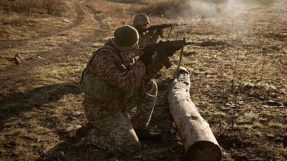 Zwei Soldaten knien hinter einem Baumstamm und schießen mit ihren Maschinengewehren.