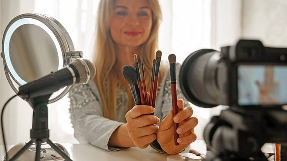 Eine Beauty-Influencerin hält verschiedene Schminkutensilien in eine Kamera