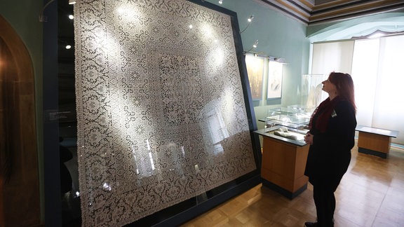 Eine Mitarbeiterin betrachtet eine Klassische Plauener Wickeldecke von 1935 im Plauener Spitzenmuseum.