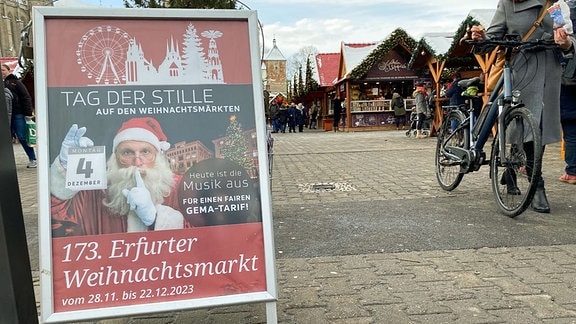 Auf dem Weihnachtsmarkt in Erfurt steht ein Plakataufsteller, auf dem ein Weihnachtsmann abgebildet ist, der sich einen Finger vor den Mund hält und so auf einen Weiohnachtsmarkt ohne Musik hinweist.