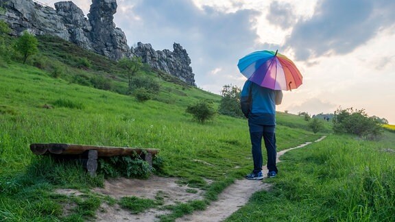 Ein Wanderer steht an der Teufelsmauer im Harz. Er hat einen Schirm in den Farben eines Regenbogens aufgespannt. 