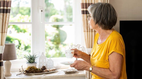 Eine ältere Frau steht in einer Wohnung allein am Fenster und trinkt eine Tasse Kaffee (gestellte Szene).