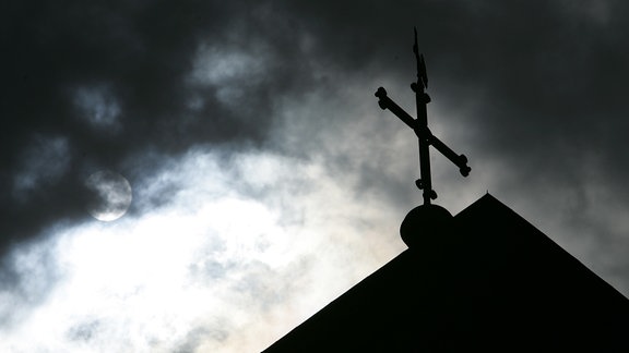 Im Gegenlicht und vor wolkenverhangenem Himmel ist eine Kirchturmspitze mit Kreuz zu sehen
