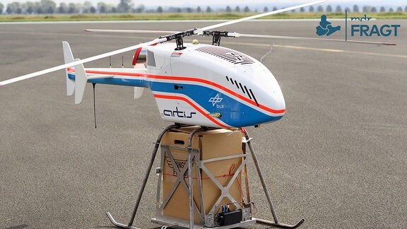 Ein Paket ist auf dem Flugfeld des Nationalen Erprobungszentrums für unbemannte Luftfahrtsysteme vom Deutschen Zentrum für Luft- und Raumfahrt (DLR) unter einer Drohne zu sehen, die das Paket tragen soll.