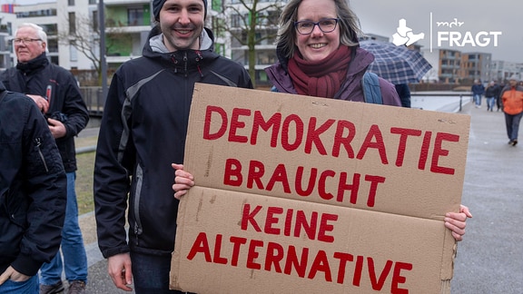 Bei einer Demo hält ein Duo ein Plakat mit "Demokratie braucht keine Alternative" hoch