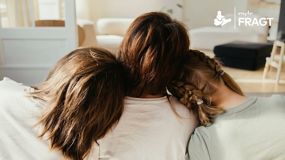 Geschwister lehnen die Köpfe auf der Schulter der Mutter im Wohnzimmer.