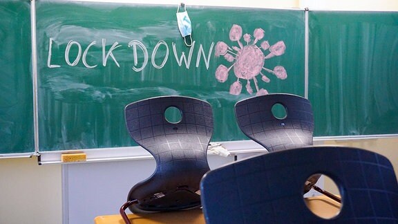 An eine Schultafel in einem Klassenzimmer wurde mit Kreide das Wort Lockdown geschrieben und ein Virussymbol gemalt. An der Tafel hängt eine Mund-Nasenmaske. Im Vordergrund sind die Stühle auf die Tische gestellt. 