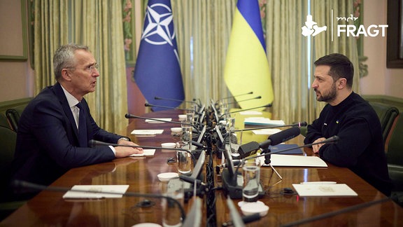 An einem Konferenztisch sitzen sich Nato-Generalsekretär Jens Stoltenberg und der ukrainische Präsident Wolodymyr gegenüber. Im Hintergrund sind die Flaggen der Nato und der Ukraine zu sehen.
