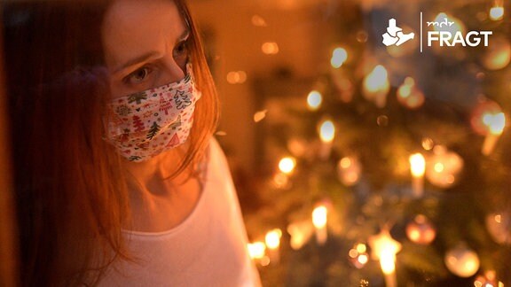 Eine Frau mit Mund-Nasenschutz-Maske schaut sehnsüchtig aus dem Wohnzimmerfenster