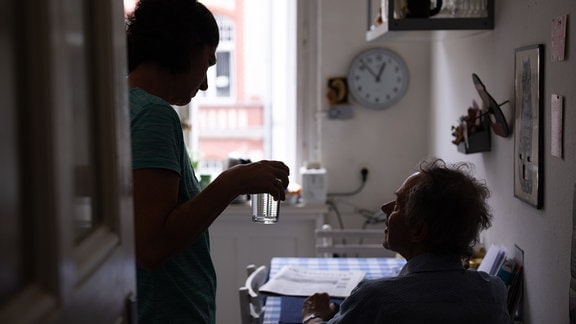 Eine Pflegerin reicht einem pflegebedürftigen Patienten bei einem Hausbesuch ein Glas Wasser.