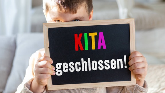 Ein kleiner Junge hält ein Schild mit Aufschrift: Kita geschlossen! 