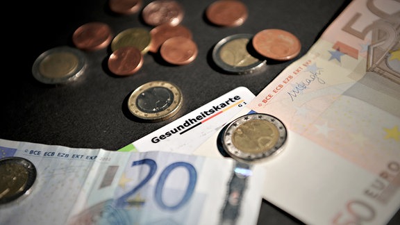 Ein Teil einer Gesundheitskarte ist zwischen Euro-Noten und Münzen zu sehen.