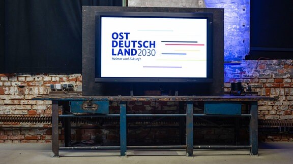 Ein Monitor mit dem Logo der Konferenz "Ostdeutschland 2030" steht auf einer Werkbank im Kunstfraftwerk in Leipzig.