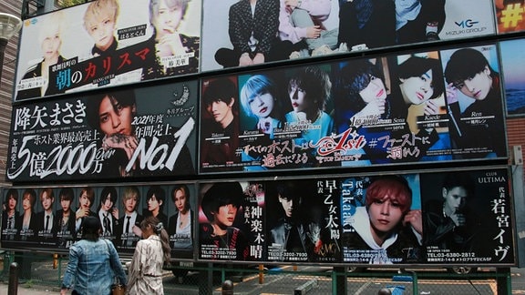 Passant*innen laufen an einer Werbewand mit mehreren Plakaten entlang.