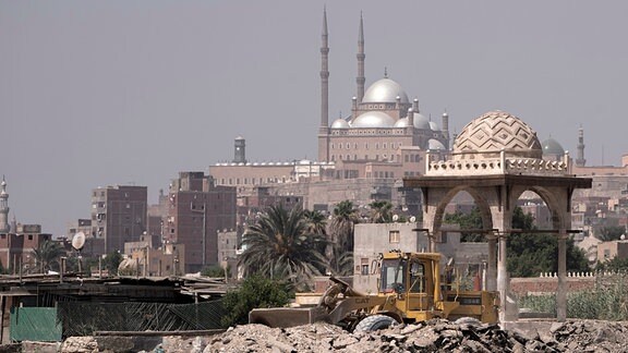 Ein Bulldozer rangiert inmitten von Trümmern vor dem Hintergrund einer Moschee.