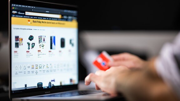 Eine Person mit einer Bankkarte in der Hand sitzt am Laptop und schaut sich die Black Friday Angebote auf der Online-Shopping-Seite von Amazon an. 