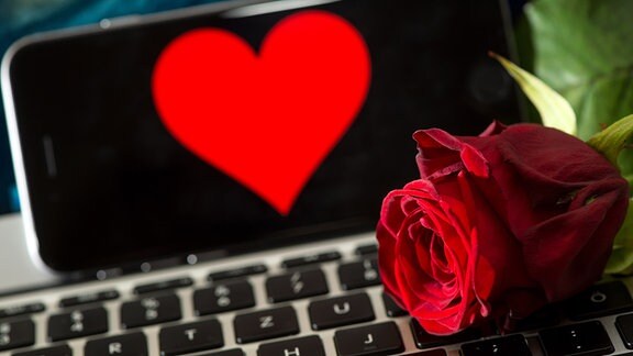 Illustration - Eine Rose liegt auf einer Computertastatur. Im Hintergrund ist ein Smartphone zu sehen, auf dem ein Herz abgebildet ist. 