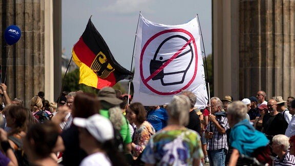 Demonstrierende halten ein Transparent mit einem durchgestichenen Emblem eines Gesichts das eine Maske trägt hoch.