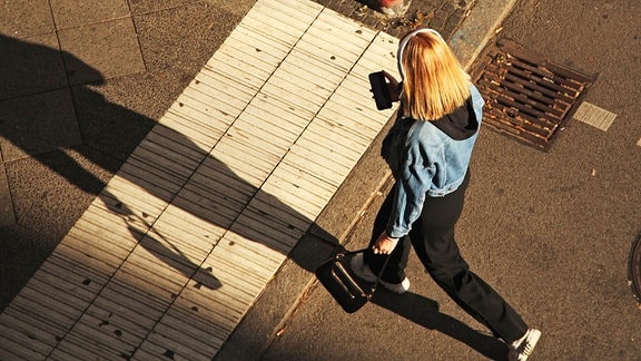 Eine junge Frau trägt Kopfhörer und blickt auf ihr Smartphone, während sie über eine Straße geht
