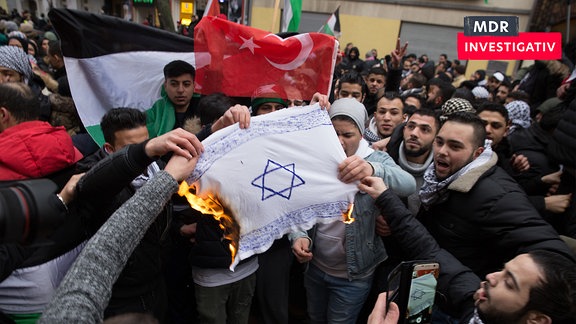 Männer verbrennen 2017 in Berlin Neukölln eine selbst gemalte Israel-Fahne. Im Hintergund ist die türkische und die palästinensische Flagge zu sehen. Einige tragen Kefia.