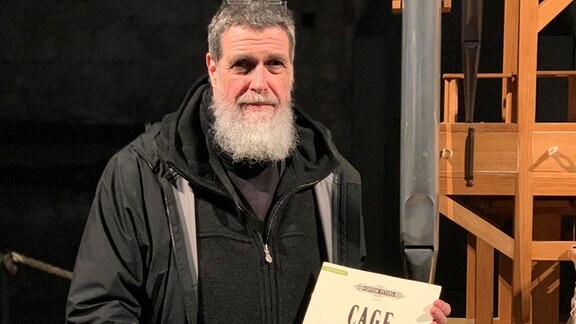 Rainer Neugebauer, ein Mann mit weißem Bart hält eine Partitur von John Cage in der Hand.