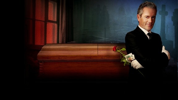 Schauspieler auf Symbolbild: Ein Mann im schwarzen Anzug steht vor einem Sarg, er hält eine Rose und Geld in der Hand, er trägt weiße Handschuhe. Im Hintergrund ist eine Friedhofsszene angedeutet