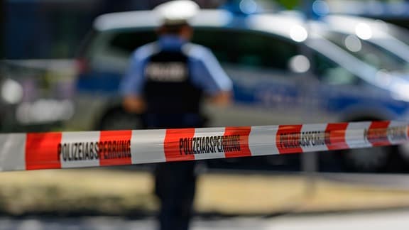 Polizeiabsperrung mit Flatterband, hinten Streifenwagen und Polizeibeamter