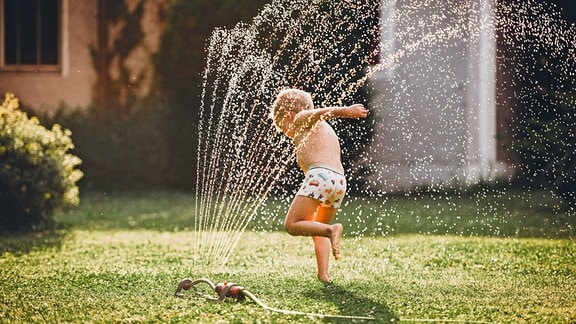 Ein Kindläuft durch einen Wasserstrahl eines Rasensprengers.