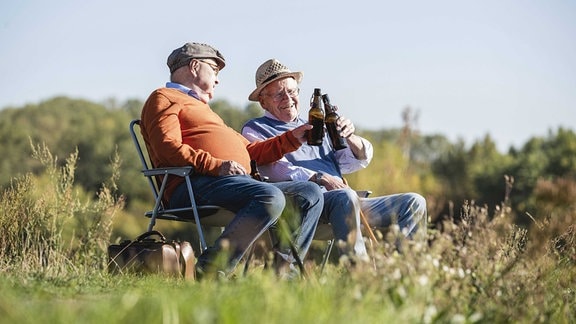Zwei Senioren sitzen auf Campingstühlen und stoßen mit Flaschen an