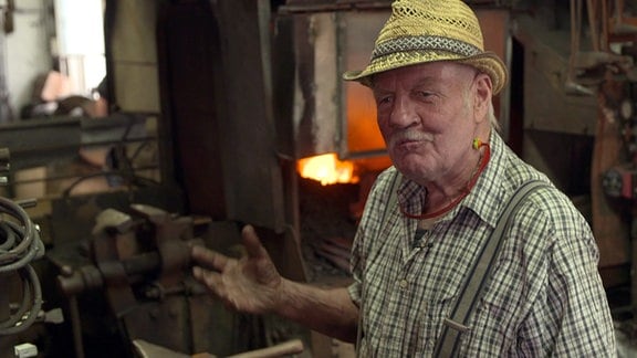 Ein älterer Mann mit Hut steht lächelnd in Werkstatt. 