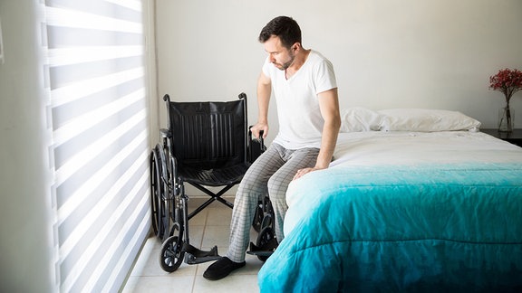 Junger Mann stützt sich vom Bett ab um sich in einen Rollstuhl zu setzten.