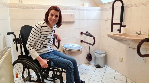 Eine Frau im Rollstuhl zeigt das behindertengerechte WC in einer Arztpraxis.