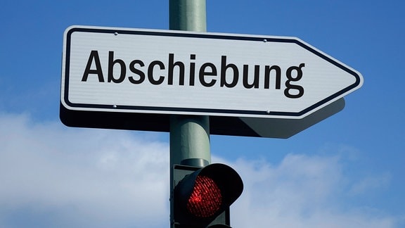 Über einer roten Ampel befindet sich ein Wegweiser mit dem Schriftzug Abschiebung