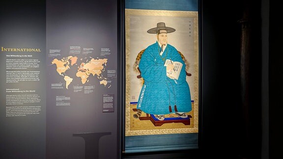Texte zu einer Ausstellung stehen in weißen lettern auf schwarzem Hintergrund. Außerdem ist eine Weltkarte abgebildet und ein Bild von einem Gelehrten in blauem Gewand und mit traditioneller asiatischer Kopfbedeckung.