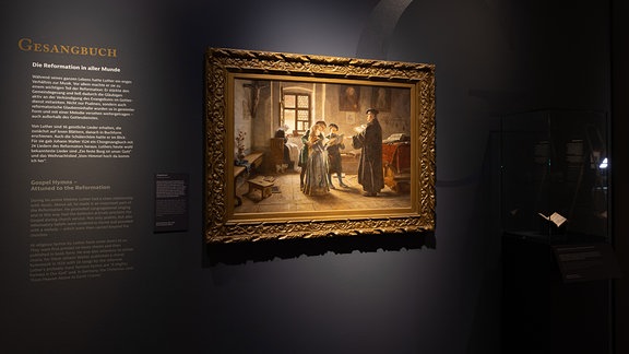 Auf schwarzem Hintergrund ist ein historisches Gemälde ausgestellt, daneben ein Ausstellungstext in weißen lettern.