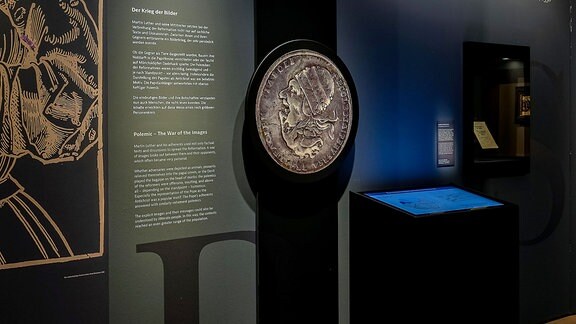 In einer Ausstellung mit schwarzen Wänden und Ausstellungstexten in weißen Lettern ist eine überdimensionale Silbermünze mit einem menschlichen Kopf im Profil zu sehen.