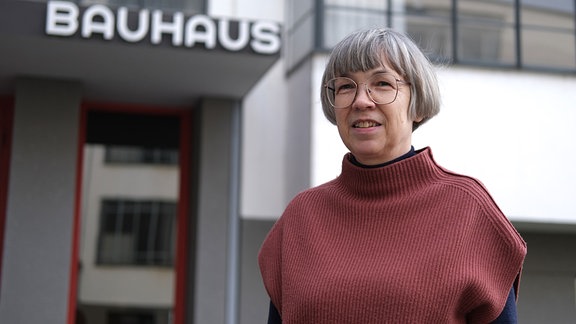 Direktorin Barbara Steiner, eine Frau mit Brille und kurzen, grauen Haaren und rotem Pullover, steht vor dem Gebäude der Bauhausstiftung in Dessau. Im Hintergrund ist der Bauhaus-Schriftzug zu erkennen.