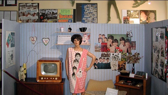 Ein nachgestaltetes Zimmer im Beatles Museum Halle, mit Postern an der Wand, einer Standpuppe mit langem T-shirt, auf dem die Beatles-Köpfe zu sehen sind. Auf einem Tisch ein sehr alter Röhrenfernseher